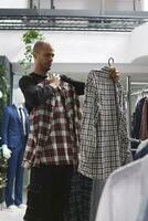 pensativo árabe hombre examinando dos camisas antes de haciendo compra decisión en ropa almacenar. compras centrar boutique comprador comprobación vestir estilo y Talla mientras elegir atuendo foto