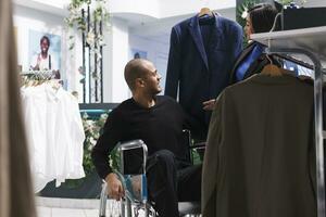 sonriente árabe hombre comprador con invalidez examinando de moda chaqueta estilo mientras vendedor participación percha con vestir en ropa almacenar. boutique empleado y cliente en silla de ruedas elegir atuendo juntos foto