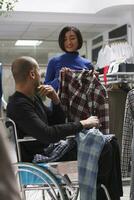 Clothing store asian woman seller helping arab man client in wheelchair to explore casual apparel. Shopping center boutique employee showing plaid shirt to customer with physical disability photo