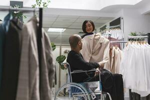 Clothing store smiling asian woman consultant helping arab man with disability in exploring formal womenswear rack. Seller assisting buyer in wheelchair in selecting beige jacket photo