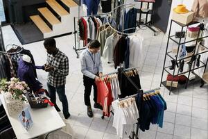 Asian employee preparing boutique for opening, arranging trendy merchandise on hangers in shopping centre. Man wearing formal shirt looking at racks full with new fashion collection in clothing store photo