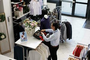 asiático trabajador preparando Tienda para apertura, arreglando de moda mercancías en perchas en moderno boutique. elegante hombre mirando a bastidores lleno con nuevo Moda colección en ropa Tienda foto