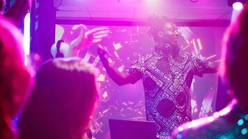 Male DJ using audio mixing station at party, putting modern music for crowd of people on dance floor. Young persons having fun partying with stage lights and live performance. Handheld shot. photo