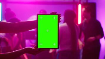Woman holding greenscreen tablet at club and showing isolated display during dance party. Young funky adult using chroma key mockup template with blank copyspace on device screen. photo