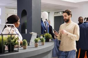 Young man customer using mobile loyalty program app while standing at checkout cashier counter in clothing store. Shopper buying clothes in shopping mall paying with smartphone photo