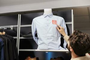 Male customer checking price on shirt while standing in clothing store, looking at red sale tag sticker. Man shopper counting discount while making purchases in clothes store on Black Friday photo