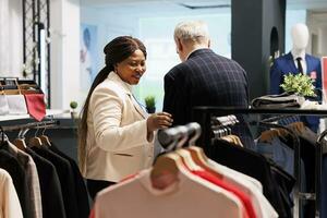 Friendly smiling black woman shop assistant helping senior man customer to choose clothes in fashion boutique. Helpful pleasant African American saleswoman serving assisting shopper in clothing store photo