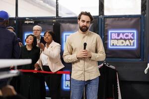 Young man reporter making TV reportage at local shopping mall during Black Friday sales, talking into microphone reporting on shopping madness in stores and how people behaving during seasonal sales photo