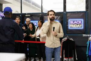 Reporter broadcasting about Black Friday madness from shopping mall. Young man TV journalist holding microphone making live reportage while standing against angry mad shoppers waiting for sales photo