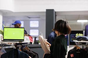 Open laptop with blank green screen in shopping mall, computer pc with mock up chroma key display standing on garment rack in fashion boutique. Woman shopper buying clothes during sales photo