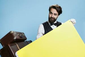 Classy bellhop shows yellow banner against blue background, stylish porter in suit and tie does ad for marketing campaign with blank poster sign. Doorman with empty billboard. photo