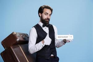 Staff member poses on camera while pointing at restaurant sign to direct visitors. Young sophisticated bellhop presents dining room indicator emblem, wall pointer symbol. photo