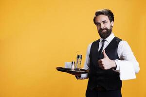 Experienced butler gives thumbs up in studio, presenting agreement symbol and being positive on camera. Young man with wiater occupation shows like approval before serving cup of coffee. photo