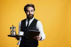 Personnel carries food bill on tablet, serving customers with cup of coffee at luxury restaurant in studio. Restaurant staff gives tab to clients on modern device, posing against yellow background. photo
