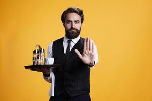 Expert waiter raising palm to do stop sign in studio, expressing disapproval and rejection on camera. Confident stylish waiter showing no symbo with arm, refusing and dismissing something. photo