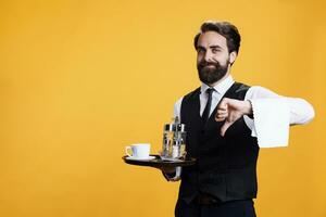 Fine dining worker shows dislike sign on camera, giving thumbs down against yellow background. Professional elegant butler presents disapproval or disagreement symbol, holds restaurant tray. photo