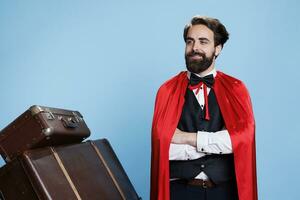 Hotel porter acting as superhero with cape, offering to help guests with trolley bags on camera. Young man bellhop presenting comic mascot with red mantle, feeling powerful and confident. photo