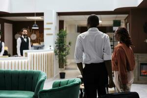 People arriving at luxury hotel lobby, entering lounge area and preparing to do check in process. Young couple talking to front desk employees about room reservation, reception counter. photo