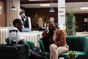 Happy people relaxing in lounge area before doing check in procedure to receive room card key, booking reservation. African american guests arriving at luxury hotel, waiting on couch. photo