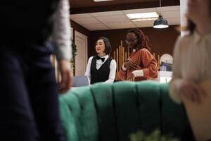 Hotel staff talking to couple in lobby, asking about room reservation booking before helping with check in procedure. Receptionist at front desk filling in registration forms, friendly service. photo