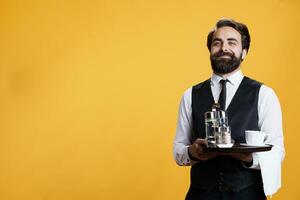 Smiling butler poses with platter in hand, standing against yellow background while he works at five star restaurant. Experienced employee with tray and towel taking orders, serving customers. photo