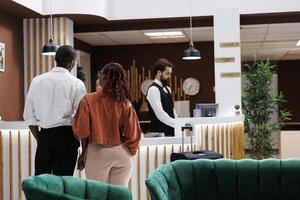 Travelling couple arriving at hotel reception desk, holding hands in lobby feeling happy about honeymoon. Guests approaching counter to do check in procedure and register at modern resort. photo