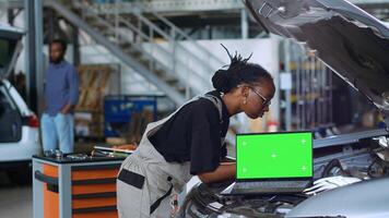 Chroma key laptop in garage workplace sitting on broken automobile. Mockup device in car service next to african american mechanics in background refurbishing damaged vehicles photo