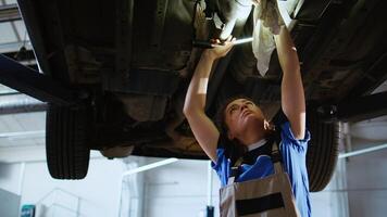 Certified technician working on suspended car in garage, checking components during routine maintenance. Auto repair shop worker walking underneath vehicle, examining it using work light photo