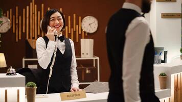 Happy smiling receptionist taking travel accommodation booking requests standing at check in desk in stylish lounge. Professional hotel employee taking phone calls while working at reception counter photo