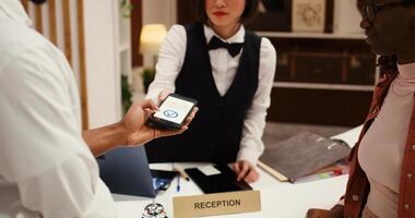 Receptionist using POS payment system to validate card purchase of extra room amenities and hotel services, close up. African american couple using smartphone banking app during booking in process photo