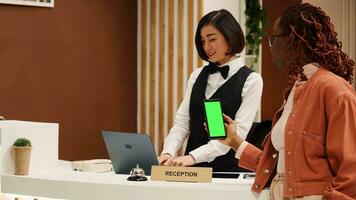Tourist holding chroma key green screen mock up smartphone waiting at front desk reception for check in process to be over. Asian receptionist processing hotel guest payment info photo