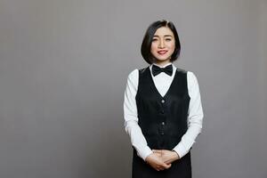 Smiling attractive asian waitress wearing uniform with bow tie standing and looking at camera. Restaurant professional receptionist posing for studio portrait on gray background photo