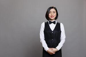 Smiling attractive asian waitress wearing uniform with bow tie standing and looking at camera. Restaurant professional receptionist posing for studio portrait on gray background photo