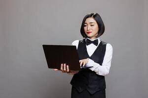 Young asian waitress using laptop, checking cafe online menu and managing catering service. Attractive restaurant receptionist wearing professional uniform and holding portable computer in studio photo