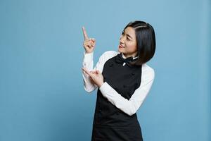 Cheerful restaurant asian waitress showing to side with fingers for service promotion. Young attractive woman receptionist in uniform pointing to right while posing in studio photo