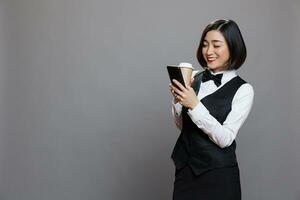sonriente asiático mujer vistiendo recepcionista uniforme mensajes de texto y desplazamiento social medios de comunicación mientras Bebiendo café a ir. restaurante alegre camarera disfrutando té en papel jarra y utilizando móvil teléfono foto