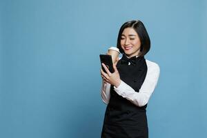 Smiling asian waitress wearing cafe uniform enjoying coffee to go and texting on smartphone. Cheerful woman receptionist holding tea paper mug and reading message on mobile phone photo