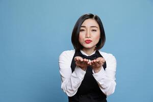 Smiling attractive asian waitress wearing uniform sending air kiss and looking at camera. Beautiful young woman receptionist expressing love and affection portrait in studio photo