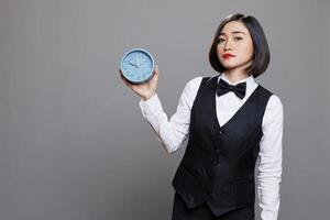 Hotel woman receptionist holding retro alarm clock and looking at camera with serious facial expression. Young asian waitress wearing uniform showing time management concept portrait photo