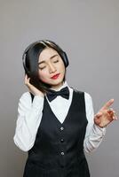 Asian woman receptionist in uniform enjoying playlist in headphones. Young attractive waitress with closed eyes listening to music in wireless earphones while posing in studio photo