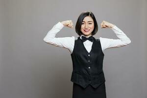 Cheerful attractive strong asian woman receptionist showcasing bicep muscles portrait. Restaurant waitress in uniform showing power and strength concept while looking at camera photo