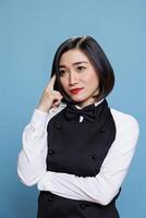 Young thoughtful aisan waitress dressed in cafeteria uniform standing in studio and contemplating solution with focused face expression. Pensive woman receptionist thinking photo