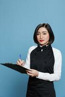 Attractive asian waitress taking notes of customer order in restaurant portrait. Young woman receptionist maintaining records, writing on clipboard with pen and looking at camera photo