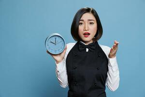Asian waitress holding alarm clock with surprised facial expression portrait. Worried young woman cafe employee wearing professional uniform showing time and looking at camera photo