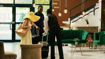 Hotel bellhop providing luxury service to guests entering reception lobby, concierge carrying baggage and trolley bags to room. Young couple arriving at tropical resort front desk. photo