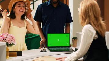 Hotel worker welcoming guests at reception, using greenscreen template on laptop at front desk. Employee talking to people in lobby, working with isolated mockup display at resort. Tipod shot. photo