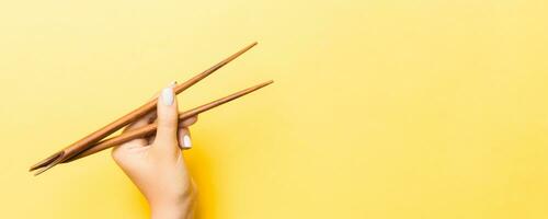 Wooden chopsticks in female hand on yellow background with empty space for your idea. Tasty food concept photo