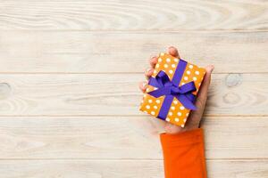 Woman arms holding gift box with colored ribbon on yellow rustic wooden table background, top view copy space photo