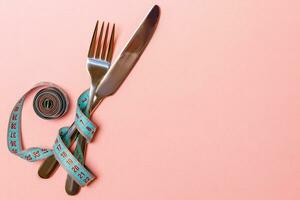 Top view of crossed knife and fork connected by measuring tape on pink background. Overeating and healthy diet concept photo