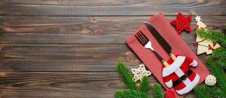 Top view of fork and knife put in santa clothes on napkin with christmas decorations and new year tree on wooden background. Banner holiday and festive concept with copy space photo